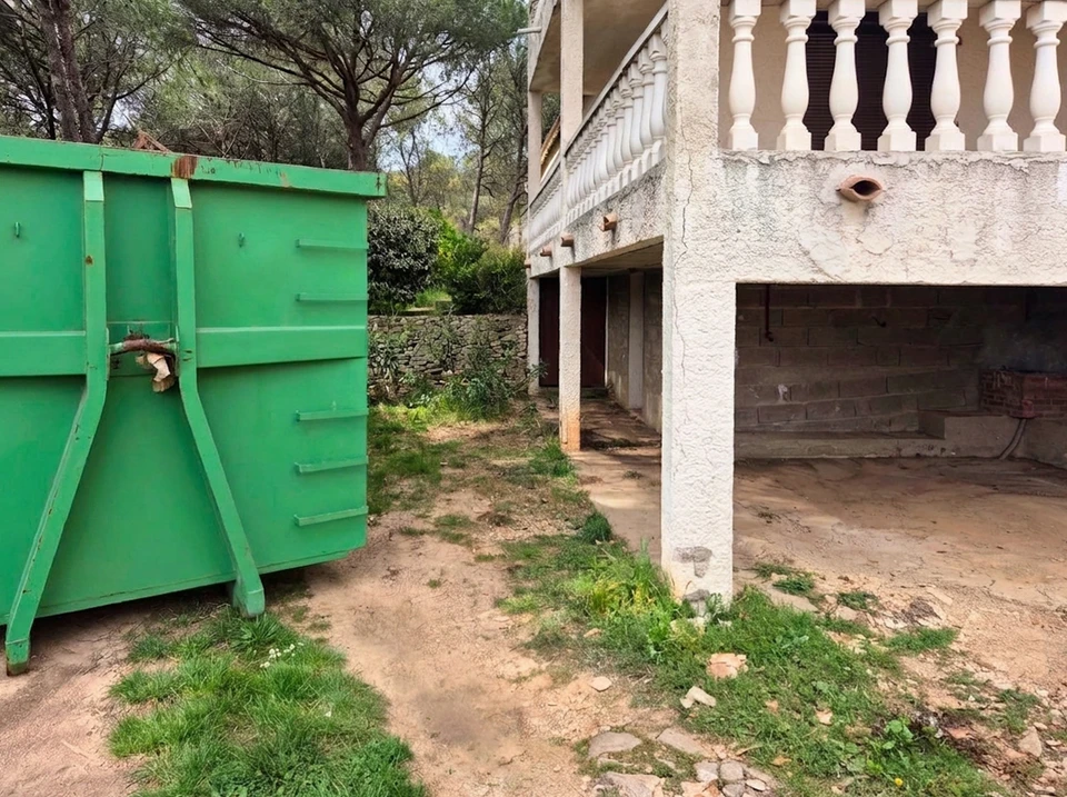 Ouvrier en tenue de travail déposant des débris dans une benne à ordures verte de grande capacité, pleine de gravats et mobilier en bois démoli
